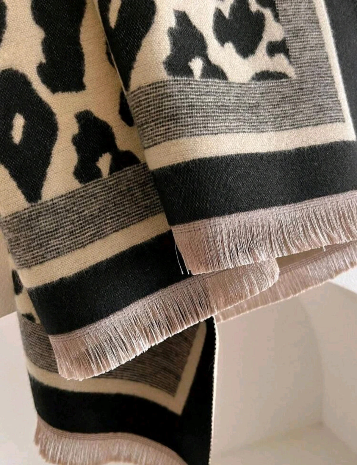 Beige scarf with black leopard print, fringed edges, reversible design, soft polyester material.